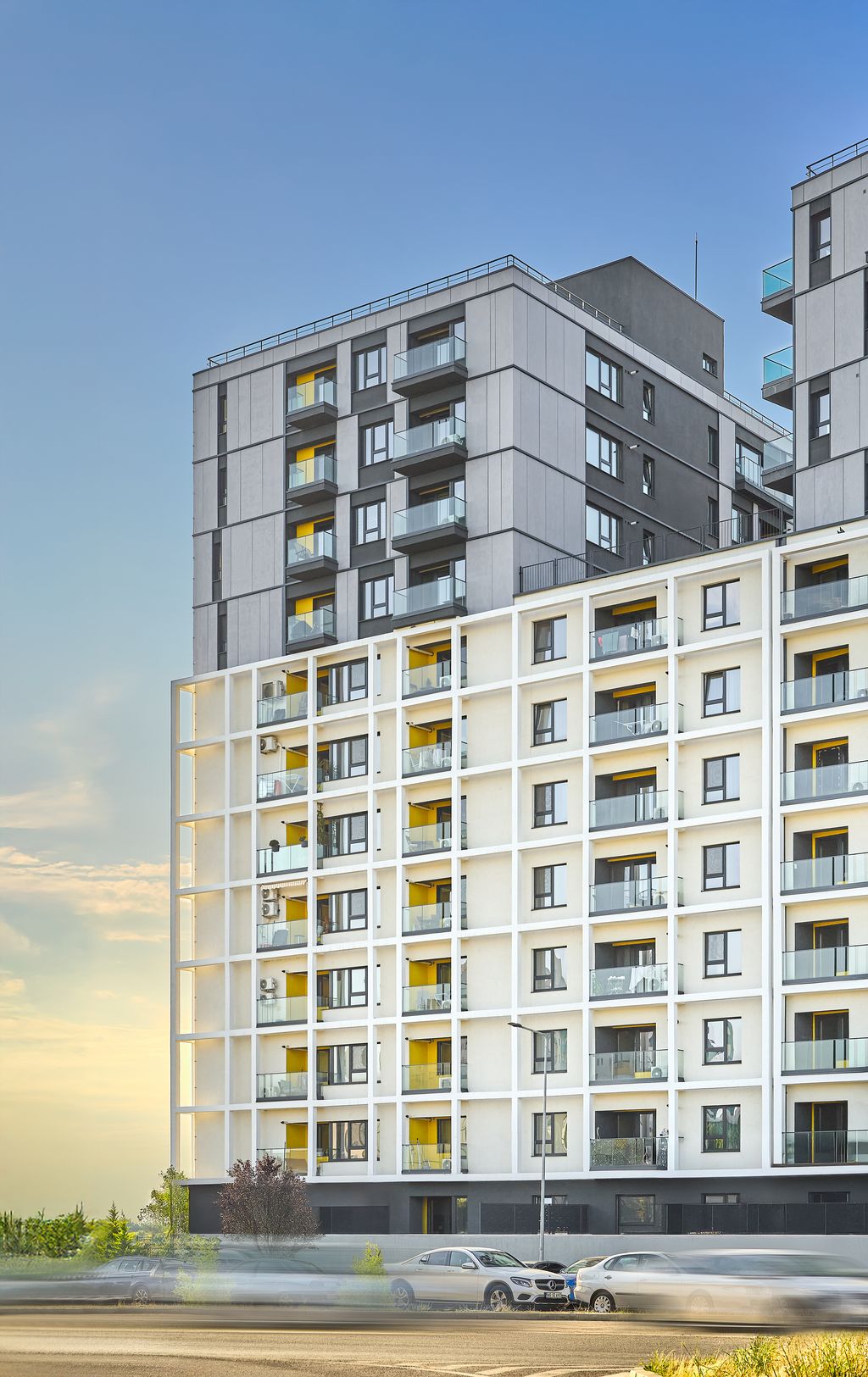 Signature street-level view in the golden evening light: in the foreground, the lower white plaster body with its white grid structure and the yellow-accented balconies; above and set back, the tall body clad in anthracite-grey panels, against a sunset sky. The hero image that clearly reads the two contrasting facade systems and the stepped massing.