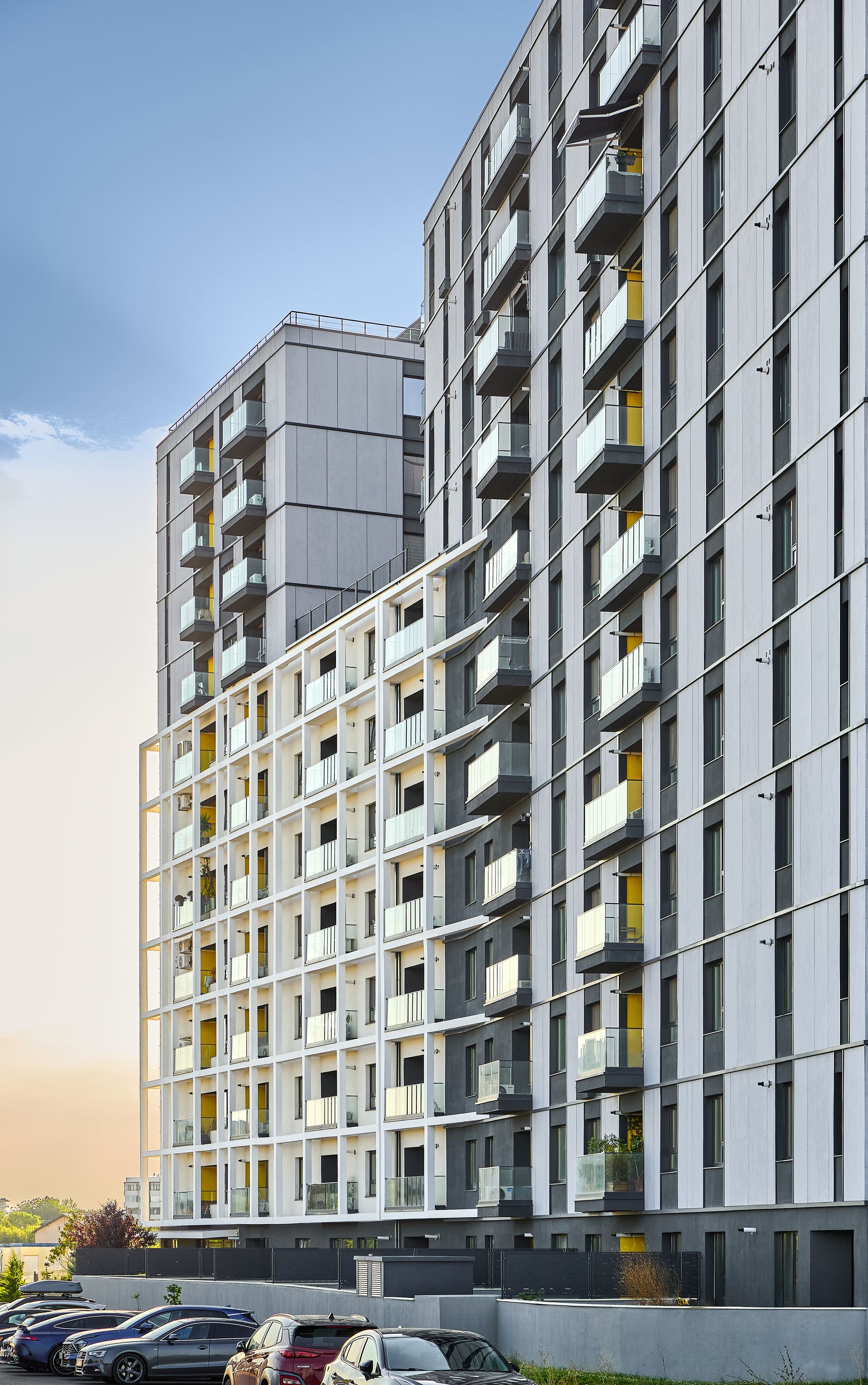Overall view of the Lake House 2 residential building facade, captured from street level in warm evening light. It reveals the fragmented, stepped massing, the anthracite-grey fibre-cement panels alternating with white plastered surfaces, the balconies with glass railings and the warm accents within the loggia recesses.