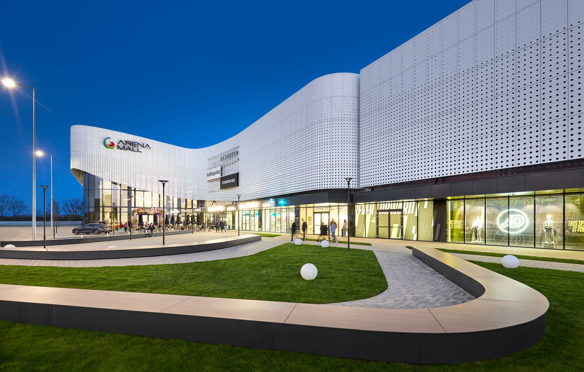 Evening-lit façade of Arena Mall Bacău, with the access pathway