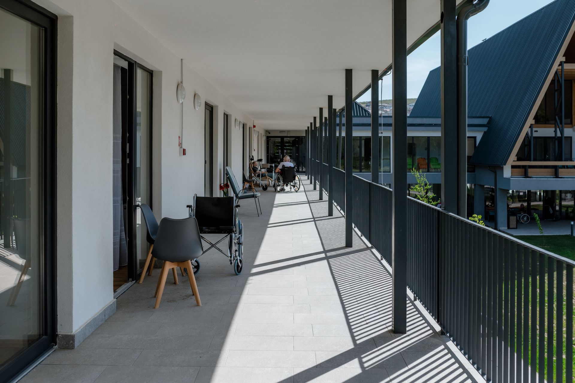 The covered exterior gallery corridor on the upper floor, with a white-plastered wall and glazed sliding doors to the rooms on one side, and metal columns and balustrade opening towards the courtyard on the other. Stone flooring; chairs and a wheelchair signal the senior-residence use, with the pitched-roof wing visible across the courtyard.