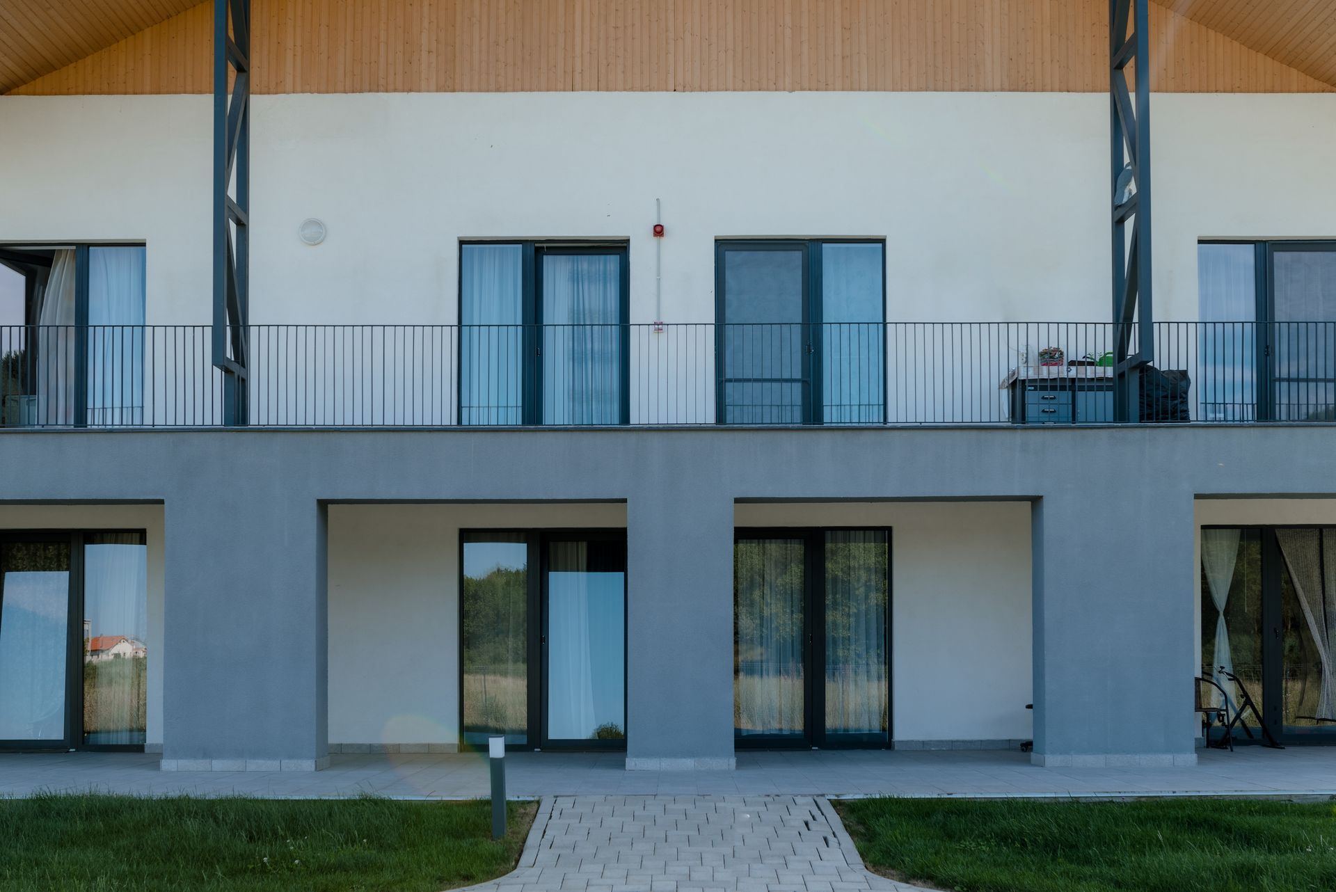 Frontal view of the residential facade: on the ground floor, recessed grey-plastered loggias/terraces with wide glazed doors; on the upper floor, a continuous balcony with a slender metal balustrade and full-height windows. Above, the timber gable of the pitched roof and the diagonal steel bracing; in the foreground, lawn and a paved path.