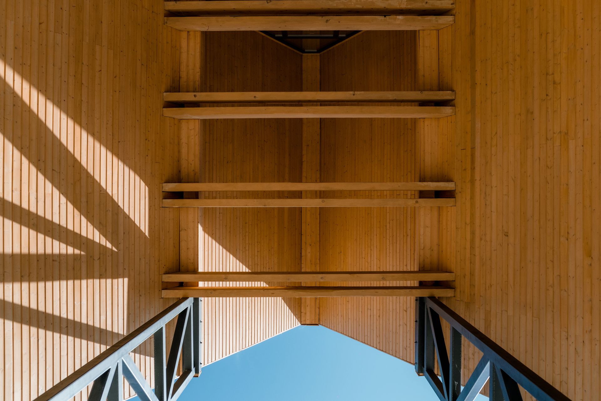 Architectural detail looking up at the roof structure: vertical natural-timber cladding and horizontal timber beams, supported by metal bracing elements, frame a triangular opening to the blue sky. The play of light and shadow emphasises the texture of the wood.