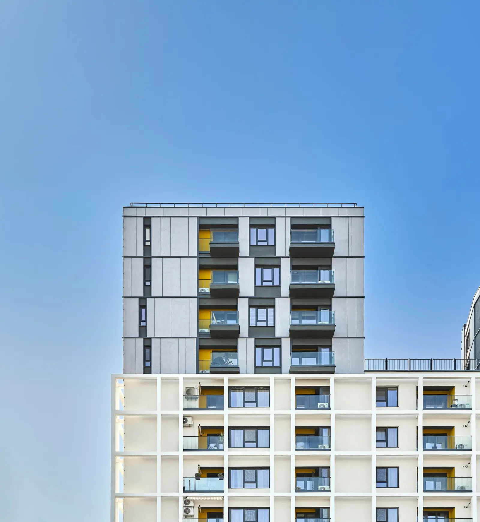 Vertical facade detail against a clear blue sky, highlighting the contrast between the two treatments: the set-back upper body clad in anthracite-grey panels with recessed dark balconies, sitting above the lower white plastered body marked by the pronounced white grid structure and the balcony recesses with yellow accents.