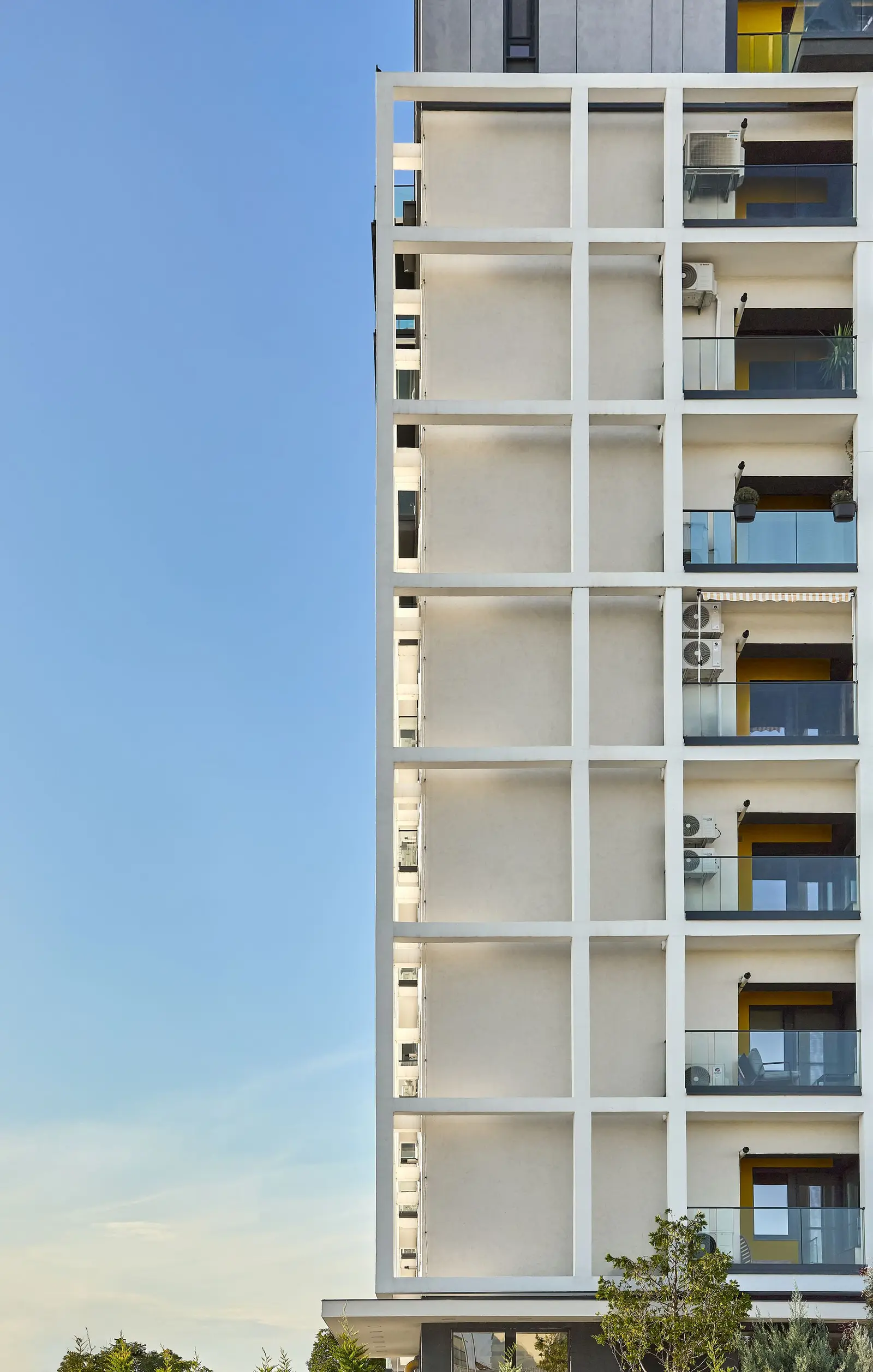 Vertical detail of the white grid facade element: the white orthogonal frame, projecting in front of the white plaster, creates a rhythm of deep loggias. Within the recesses one can glimpse interiors with yellow accents, glass railings and greenery, underlining the depth, shadows and ordered composition of this facade.