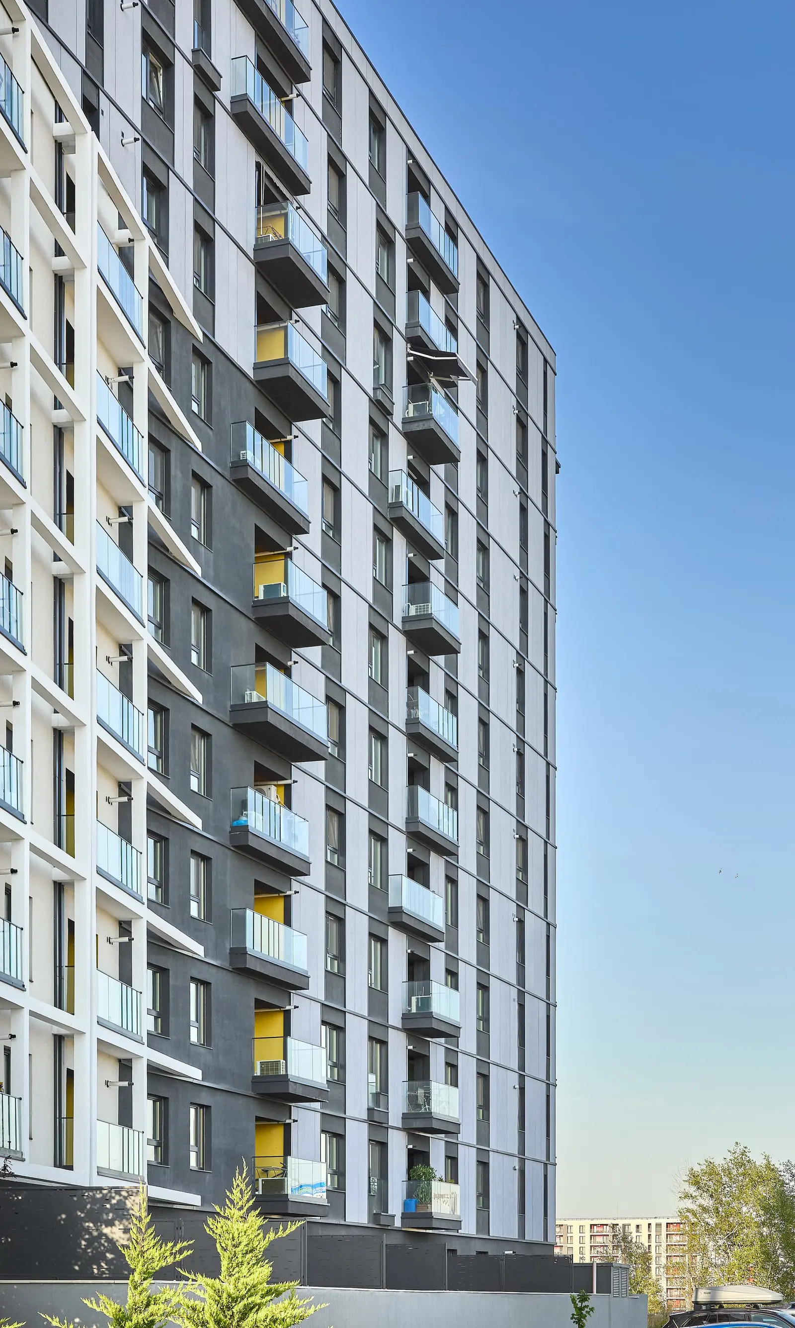 Vertical perspective view along the anthracite-grey panelled facade, taken from ground level looking upward. It reveals the rhythm of cantilevered balconies with glass railings, the fibre-cement panels with white plaster accents, the yellow-toned balcony recesses and the perspective effect that emphasises the height and the vertical registers of windows.