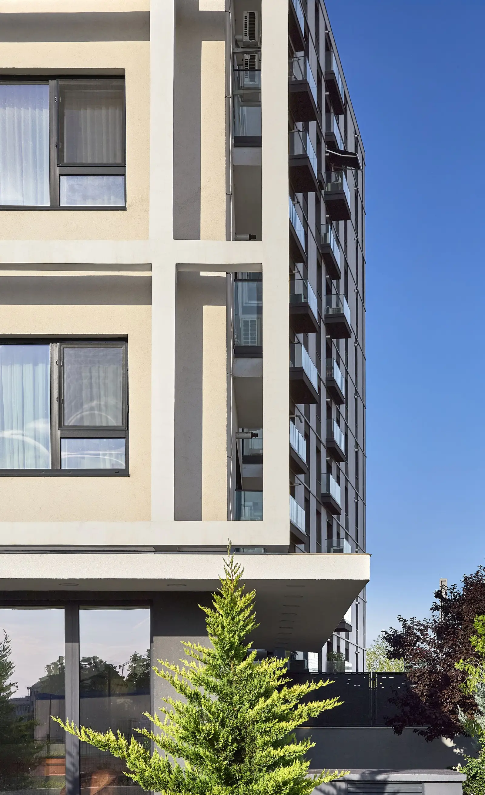 Corner detail capturing the meeting of the two facade systems: the white plaster with its grid structure (in a warm cream tone) on the left and the anthracite-grey cladding with dark balconies on the right, against a blue sky. At ground level, the glazing and a landscaped terrace with a conifer and greenery illustrate the building's relationship with the landscape.