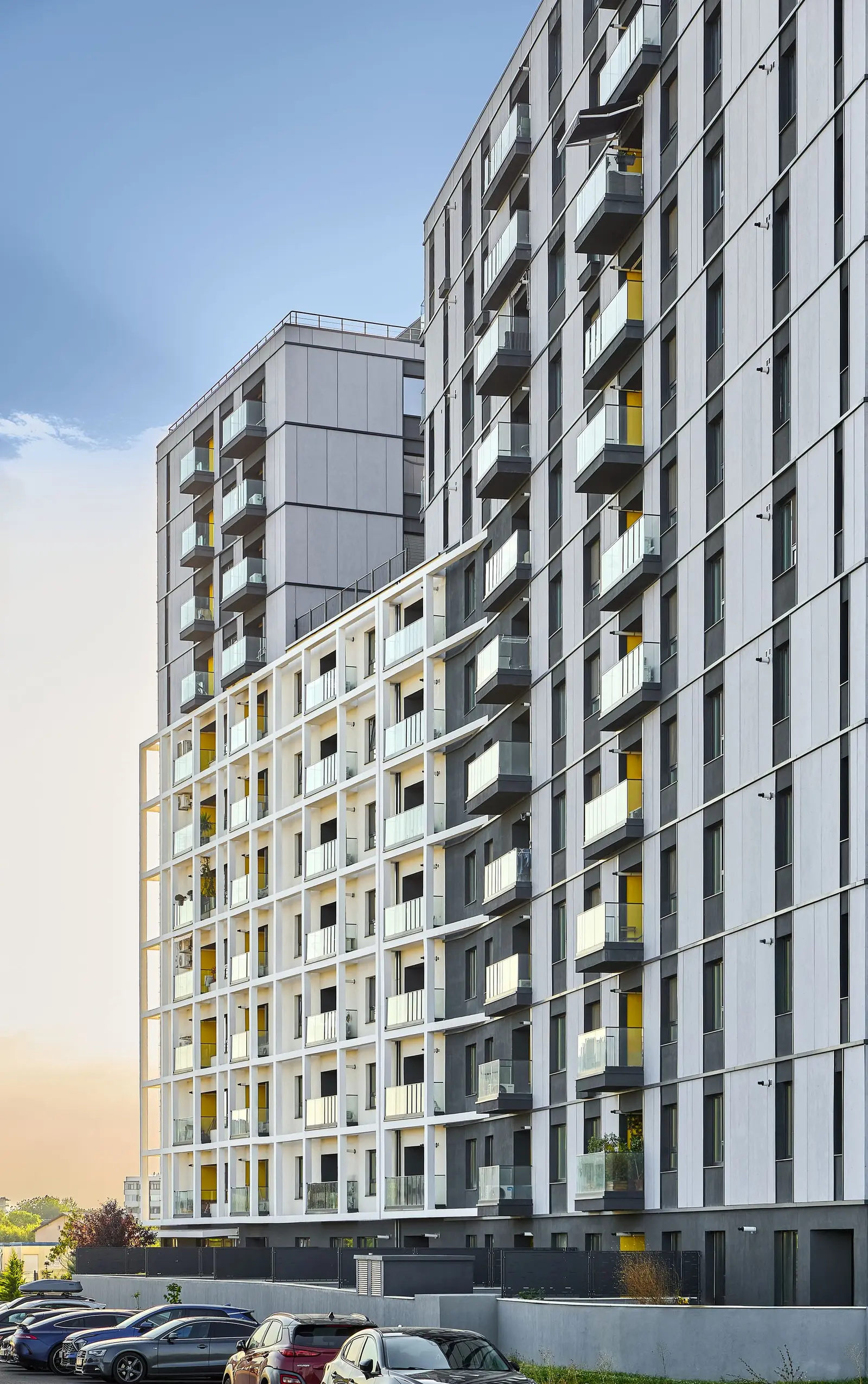 Overall view of the Lake House 2 residential building facade, captured from street level in warm evening light. It reveals the fragmented, stepped massing, the anthracite-grey fibre-cement panels alternating with white plastered surfaces, the balconies with glass railings and the warm accents within the loggia recesses.