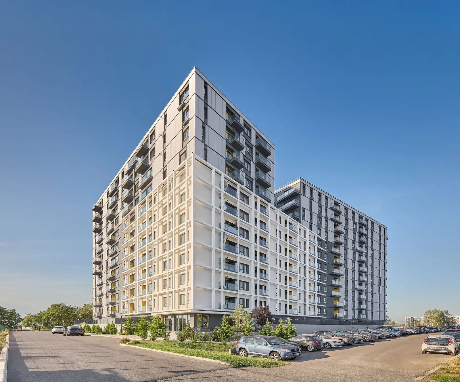 Overall three-quarter corner view of the entire Lake House 2 residential complex under a clear blue sky. It shows the fragmented volumetric composition — the tall bodies clad in anthracite-grey panels and the white plastered body marked by a white grid structure overlaid on the balconies, together with the landscaping and the parking area in the foreground.