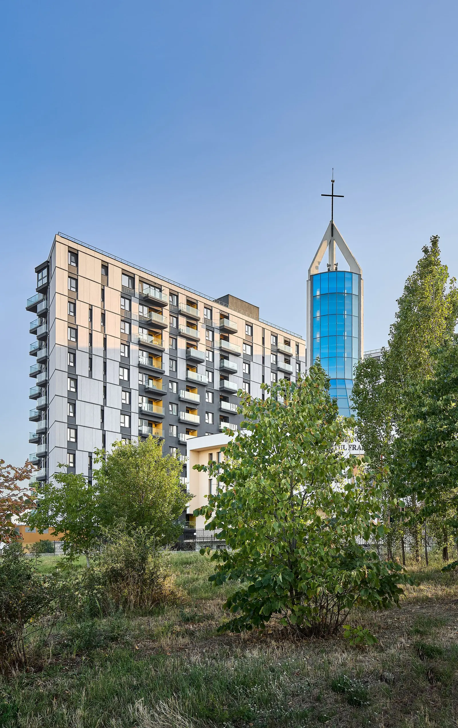 Contextual image of the building, seen from a distance across a landscaped green area with trees and shrubs in the foreground. Next to the residential body rises the glass tower of the modern church, with a blue glazed spire topped by a cross, illustrating how the complex sits within the natural and urban context of its surroundings.