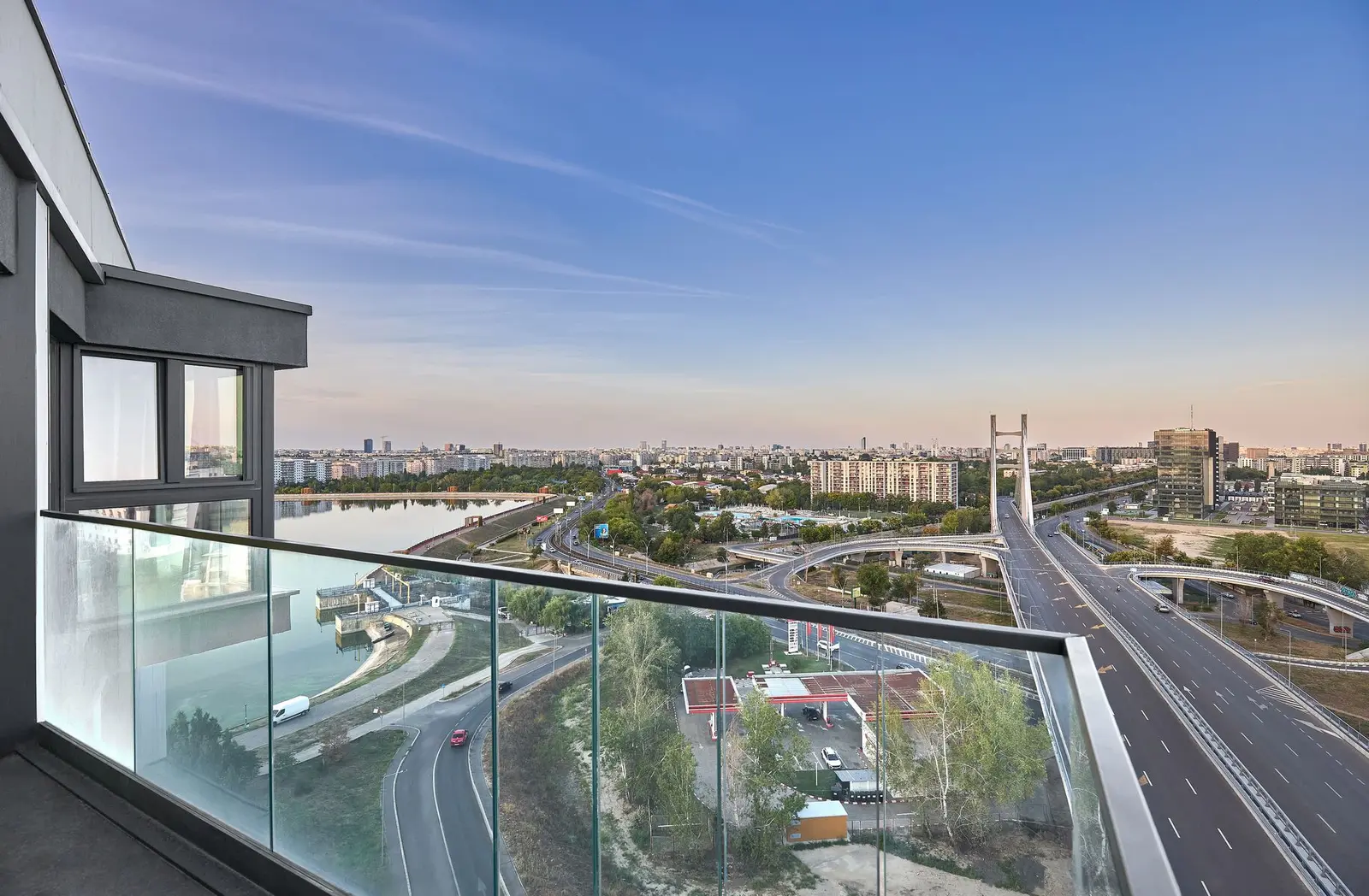 View from one of the upper balconies: the glass railing and dark facade frame the city panorama. Lacul Morii lake, the cable-stayed bridge with its distinctive pylon, the roads and interchanges, green spaces and the Bucharest skyline stretching to the horizon are all visible under a soft evening sky — an illustration of the views the apartments offer.