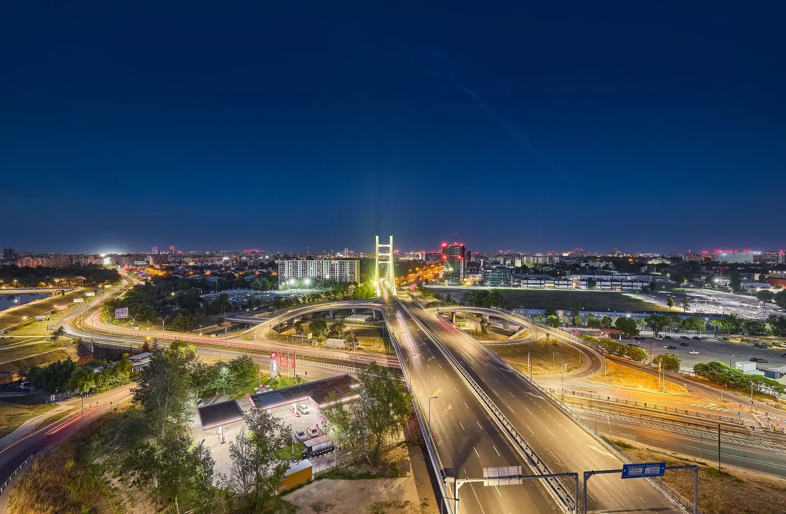 Night panorama of the surrounding area: the illuminated pylon of the cable-stayed bridge dominates the view, while the roads and interchange are traced by the light-trails of passing cars. A petrol station, green spaces and the Bucharest skyline reaching to the horizon are visible under a deep blue sky — a context image highlighting the complex's strategic location near the lake and the major traffic arteries.