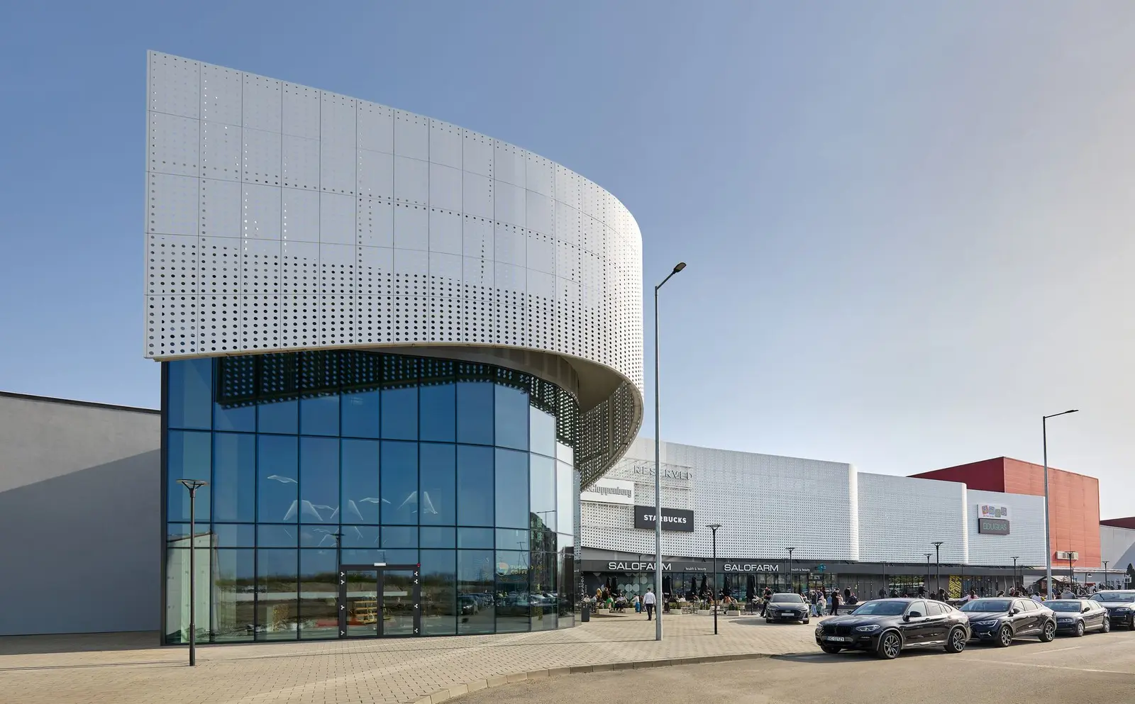 Colțul curbat al volumului alb al Arena Mall Bacău, lângă parcare