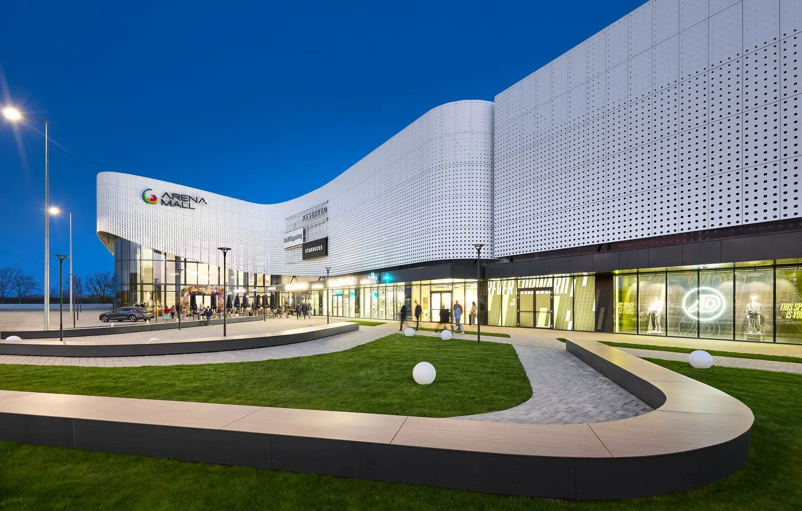 Evening-lit façade of Arena Mall Bacău, with the access pathway