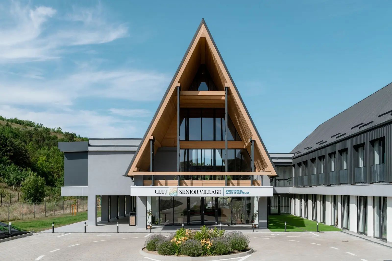 Main facade of the Cluj Senior Village complex, with the central A-frame volume, timber gable and full-height glazing carrying the community sign “Valea Izvoarelor”. In the foreground, a circular bed planted with lavender and flowers, the paved pedestrian plaza and green lawn, with the wooded hillside to the left.