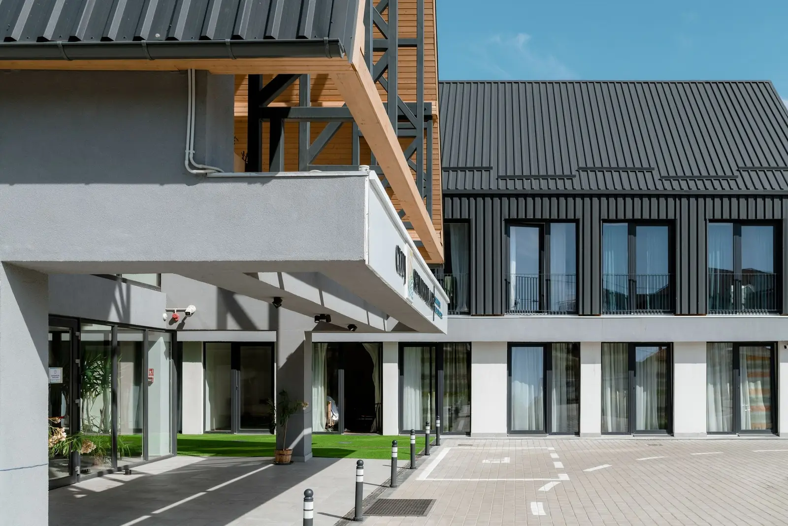 Inner courtyard of the Cluj Senior Village complex at the entrance: on the left, the grey-rendered volume with its cantilevered canopy and the timber soffit of the A-frame pavilion carried on dark steel bracing, with glazed sliding doors at ground level. On the right, the two-storey accommodation wing with a black standing-seam roof and dark vertical cladding above a white-plastered ground floor; in the foreground, the paved courtyard with bollards.