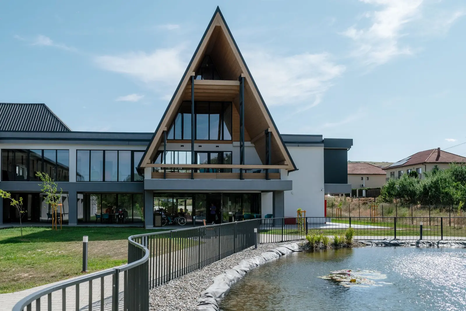 View from the garden, across the landscaped water feature, towards the central A-frame pavilion and the two-storey glazed common area. A curved metal-railed path follows the pebble-lined edge of the lily pond, set among lawns, young trees and hills in the background.