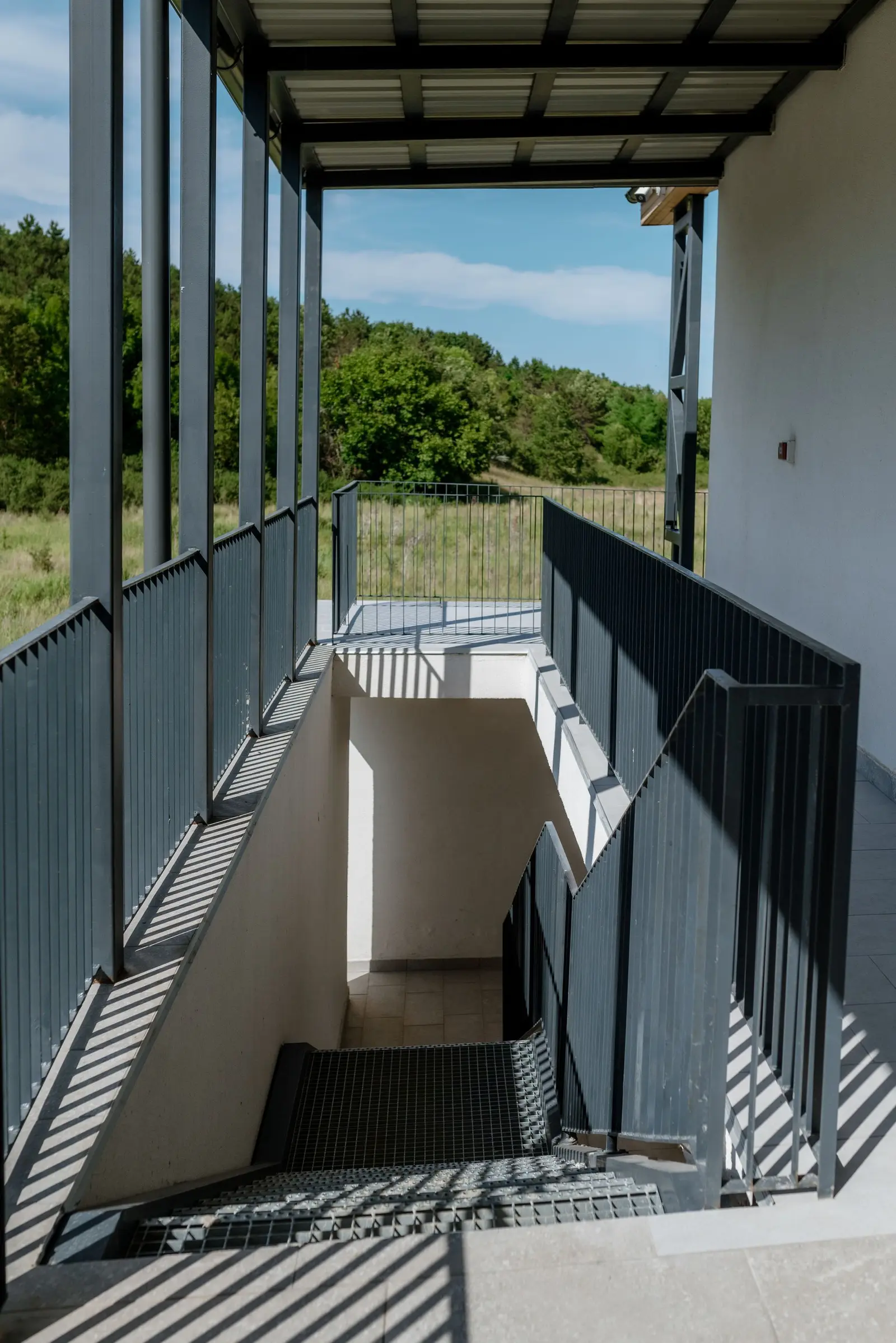 An exterior terrace/staircase sheltered by a metal pergola, with dark-metal balustrades and metal-grating treads. White-plastered walls and the lightweight structure open the view towards the natural landscape — forest and meadow — under a clear sky.