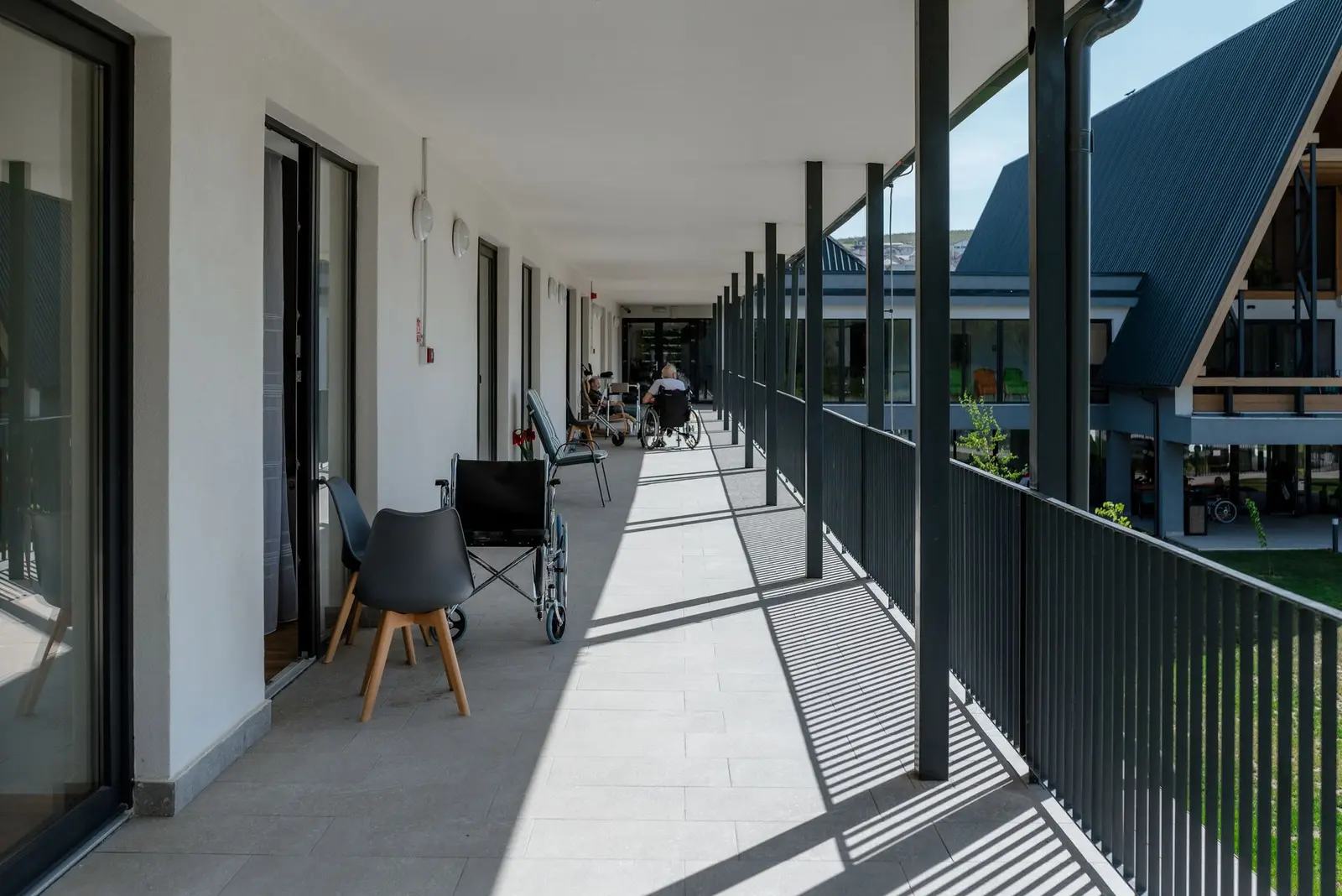 The covered exterior gallery corridor on the upper floor, with a white-plastered wall and glazed sliding doors to the rooms on one side, and metal columns and balustrade opening towards the courtyard on the other. Stone flooring; chairs and a wheelchair signal the senior-residence use, with the pitched-roof wing visible across the courtyard.