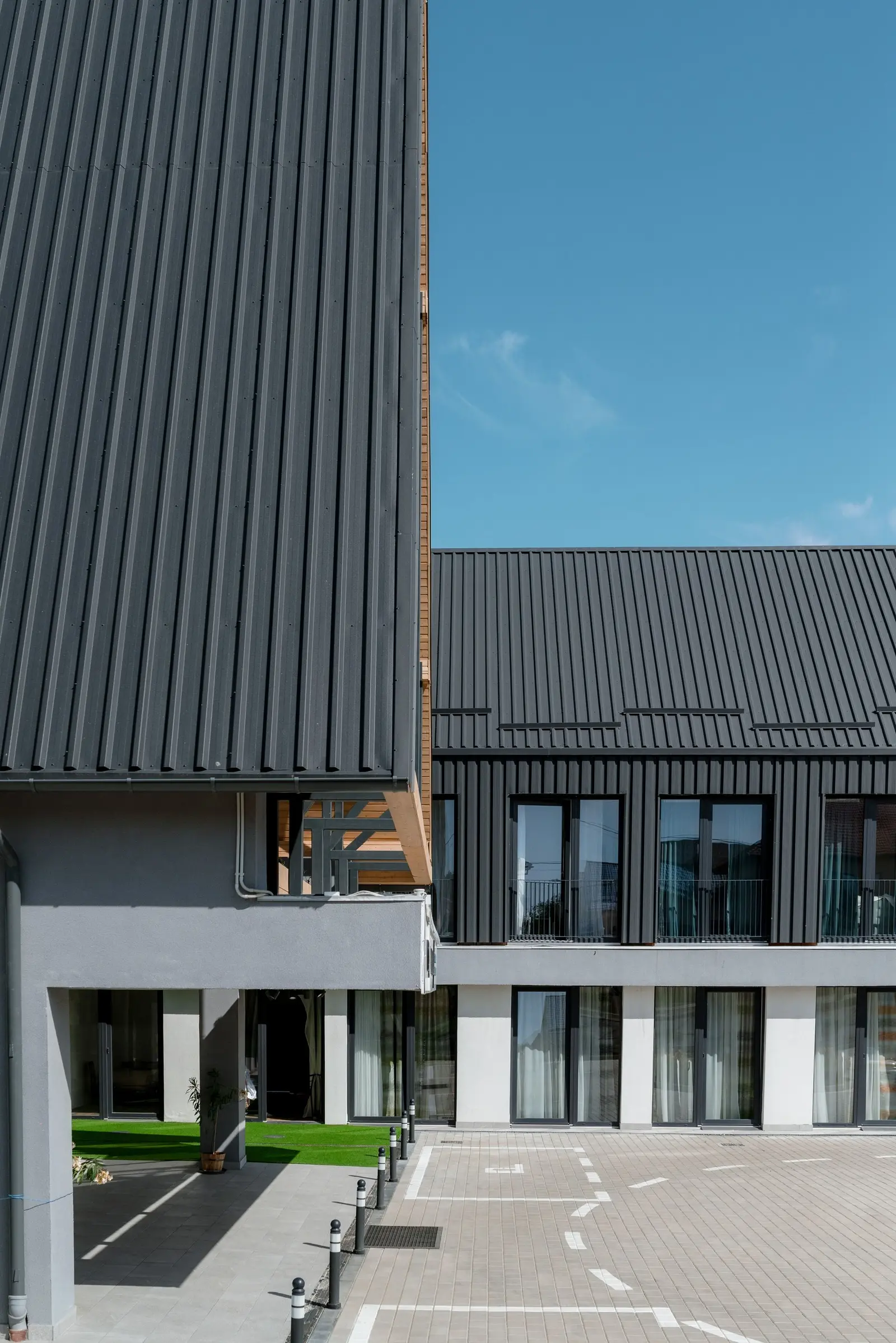 Inner courtyard of the complex: the roof and vertical cladding in dark metal contrast with the white-plastered ground floor. Note the dark-framed windows and glazed doors, the covered entrance, the paved courtyard with markings and bollards, and a small green area, under a clear sky.