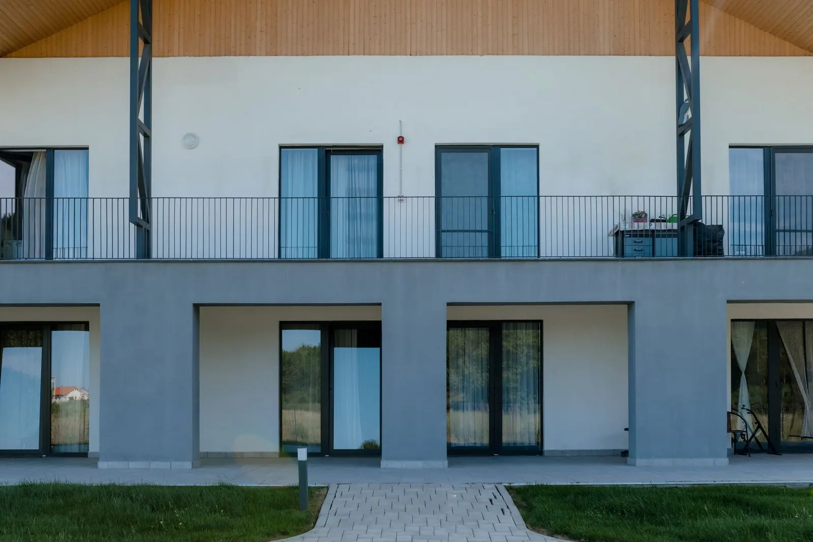 Frontal view of the residential facade: on the ground floor, recessed grey-plastered loggias/terraces with wide glazed doors; on the upper floor, a continuous balcony with a slender metal balustrade and full-height windows. Above, the timber gable of the pitched roof and the diagonal steel bracing; in the foreground, lawn and a paved path.