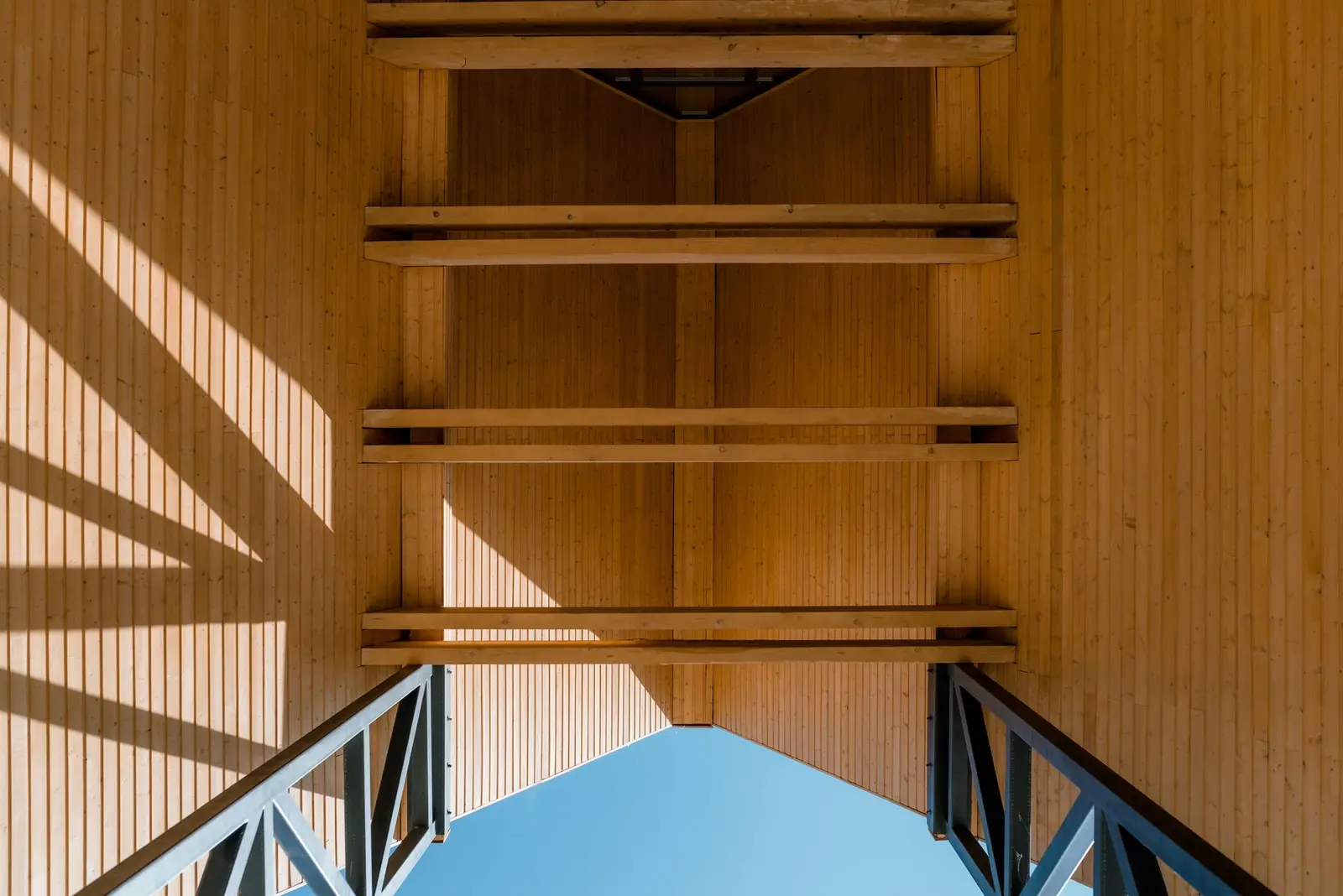 Architectural detail looking up at the roof structure: vertical natural-timber cladding and horizontal timber beams, supported by metal bracing elements, frame a triangular opening to the blue sky. The play of light and shadow emphasises the texture of the wood.