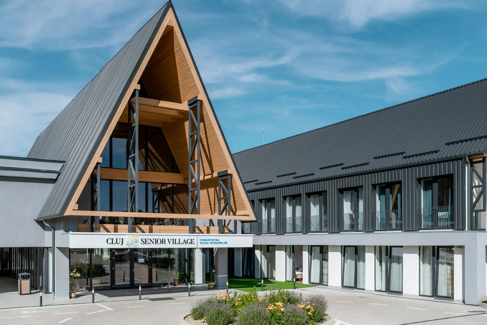 Overall view of the main entrance and the two-storey residential wing. The A-frame landmark volume, with exposed timber structure and standing-seam metal roof, connects to the accommodation block clad in dark vertical panels, with balconies and a white-plastered ground floor; in front, the paved plaza and planted roundabout.