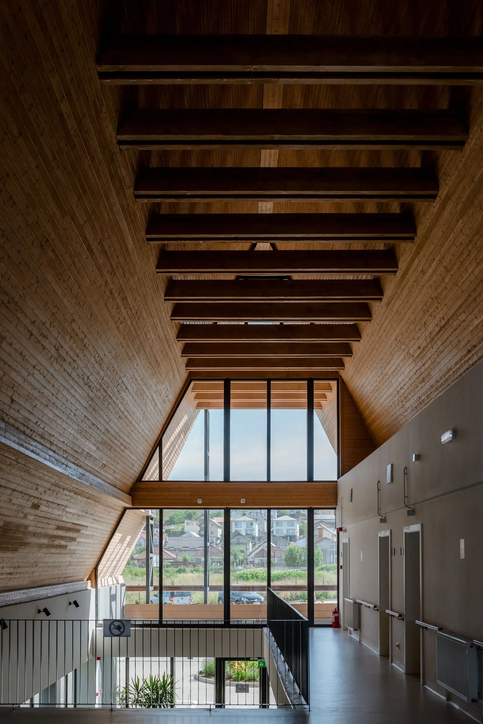 Interior of the double-height entrance hall beneath the A-frame gable: a vaulted ceiling clad in warm timber with exposed beams, and a full-height glazed curtain wall framing views of the surrounding hills. To the side, the upper-floor gallery with its balustrade and the corridor with room doors.