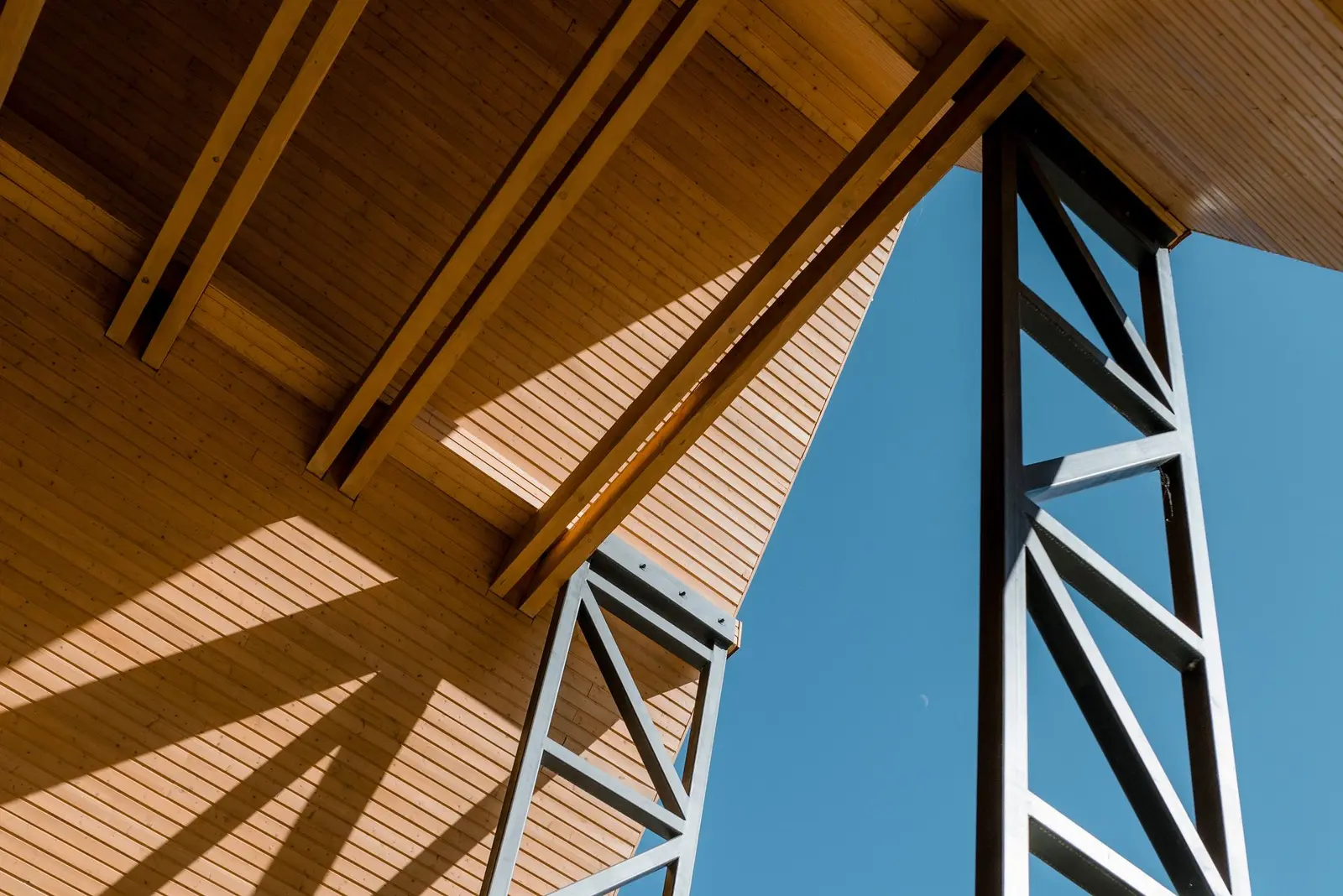 Detail of the entrance eaves: the warm timber soffit and rafters meet the slender, lattice-braced steel columns against a deep blue sky. The image highlights the dialogue between wood and steel that defines the architectural expression of the complex.
